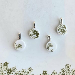 May include: Four small, round pendants with silver-colored bails. Each pendant features a white background with a green floral design. The pendants are arranged on a white plate with a green floral pattern around the edge.