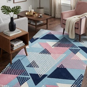 May include: A geometric patterned area rug with a light blue background and pink, white, and navy blue triangles. The rug is in a living room setting with a pink armchair, wooden furniture, and framed artwork on the wall.