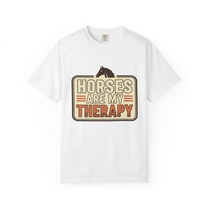 Op de afbeelding: Een wit t-shirt met korte mouwen en een rechthoekige afbeelding met een donkerbruin silhouet van een paard bovenaan. De tekst HORSES ARE MY THERAPY staat in een vetgedrukt retro lettertype met oranje en bruine accenten in een bruine rand.