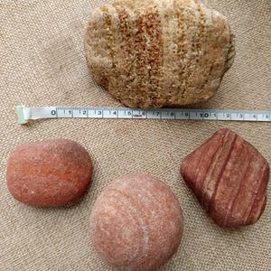 Op de afbeelding: Drie natuurlijke stenen van verschillende vormen en kleuren worden getoond met een wit meetlint. De grootste steen is beige met bruine strepen, terwijl de twee kleinere stenen roodbruin zijn, waarvan er één rond is en de andere hoekig.