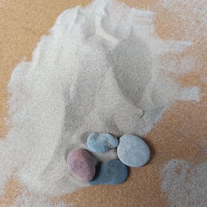 May include: A small pile of fine, light beige sand with four smooth, rounded stones resting at the base. The stones are varied in colour, including one reddish-brown, one dark charcoal grey, one light grey, and one medium blue-grey pebble.