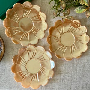 May include: Three round glass plates with a scalloped edge and a metallic peach iridescent finish. Each plate features a central circular depression surrounded by a radiating sunburst pattern of embossed lines and a textured surface.