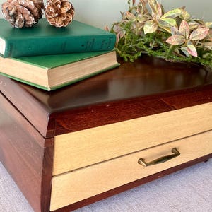 May include: A wooden desk organizer featuring a dark brown finish with a lighter natural wood drawer front and a brass-toned metal handle. The top surface is flat and smooth, providing space for two green hardcover books and pinecones.