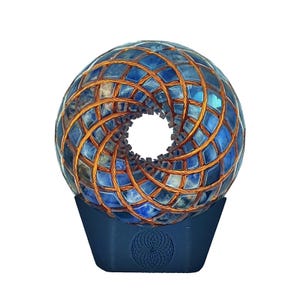 Può includere: Scultura a forma di sfera con piastrelle blu e filo di rame in un motivo geometrico. La scultura è montata su una base blu scuro con un disegno circolare. Il design complessivo è simmetrico e accattivante.