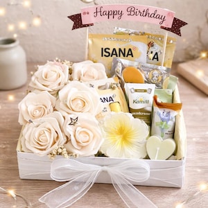Könnte beinhalten: Eine weiße Geschenkbox mit einer durchsichtigen Schleife, gefüllt mit cremefarbenen Rosen, Schönheitsprodukten und einem "Happy Birthday" Banner. Die Box enthält verschiedene Artikel, darunter eine herzförmige Kerze und eine sternförmige Dekoration.