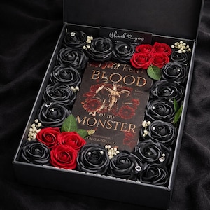 Könnte beinhalten: Eine schwarze Geschenkbox gefüllt mit schwarzen und roten Rosen, einem Buch mit dem Titel "Blood of My Monster" und einer kleinen Karte mit "thank you". Die Rosen sind um das Buch herum angeordnet.