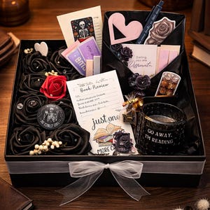 Könnte beinhalten: Schwarze Geschenkbox gefüllt mit schwarzen Satinrosen, einer roten Rose und verschiedenen Schreibwaren. Enthält eine Kerze, einen Stift, einen Lesezeichen und Karten mit Text. Eine weiße Schleife ziert die Vorderseite der Box.