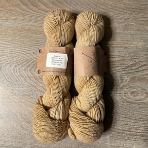 Op de afbeelding: Twee strengen lichtbruin garen, elk met een papieren etiket. De etiketten bevatten informatie over de samenstelling en het gewicht van het garen. Het garen is strak opgerold, waardoor de textuur van de vezels zichtbaar is. De achtergrond is een lichtbruin houten oppervlak.