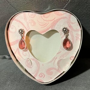 May include: A pair of silver-toned drop earrings with pink teardrop-shaped stones, displayed inside a heart-shaped gift box. The box has a pink and white floral design and a heart-shaped cutout. The earrings are suspended in front of a light green background.