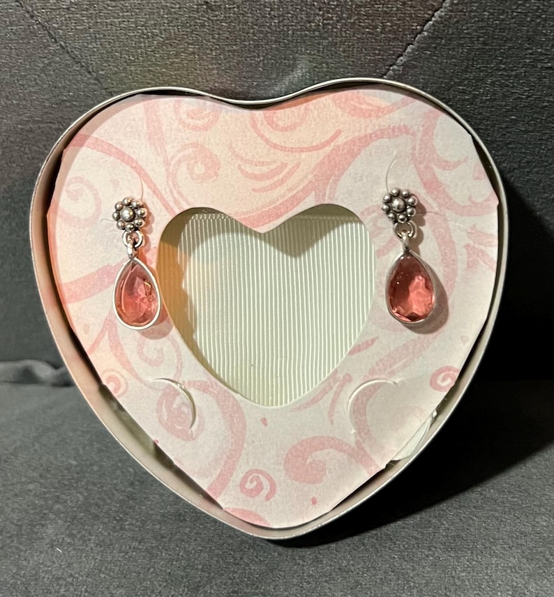 May include: A pair of silver-toned drop earrings with pink teardrop-shaped stones, displayed inside a heart-shaped gift box. The box has a pink and white floral design and a heart-shaped cutout. The earrings are suspended in front of a light green background.