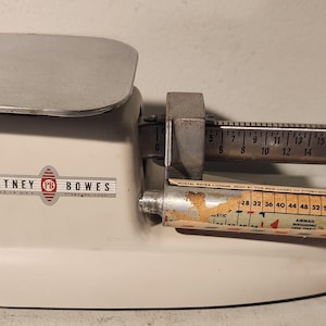 May include: White vintage Pitney Bowes postal scale with a silver weighing platform and a sliding weight bar with markings in ounces. The scale has a red and white label that reads "Pitney Bowes" and "Made in U.S.A."