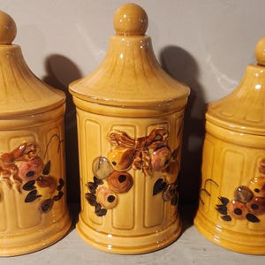 May include: Set of three ceramic canisters with a yellow glaze and floral designs. The canisters have a lid and are decorated with brown, pink, and white flowers.