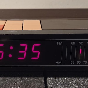 May include: A vintage Toshiba clock radio with a digital display showing 5:35 in bright pink. The radio has a brown and black casing with orange and tan buttons. The word "TOSHIBA" is printed on the front.