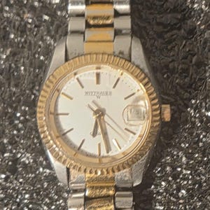 May include: A wristwatch with a silver and gold-tone metal band and a white face. The watch face has gold-tone hands and markers, with the brand name "Wittnauer" displayed. The watch band is made of alternating silver and gold-tone links.