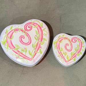 May include: Two heart-shaped decorative tins. The tins are white with a pink heart design, accented with green vines and berries. The word "Brighton" is written in pink script on the bottom right of each tin.
