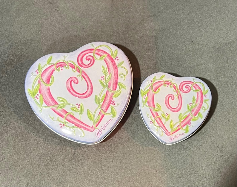 May include: Two heart-shaped decorative tins. The tins are white with a pink heart design, accented with green vines and berries. The word "Brighton" is written in pink script on the bottom right of each tin.