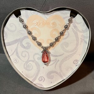 May include: A silver-toned necklace with a teardrop-shaped, pink gemstone pendant. The necklace is displayed in a heart-shaped box with the brand name "Brighton" printed on the inside. The necklace has a floral design.