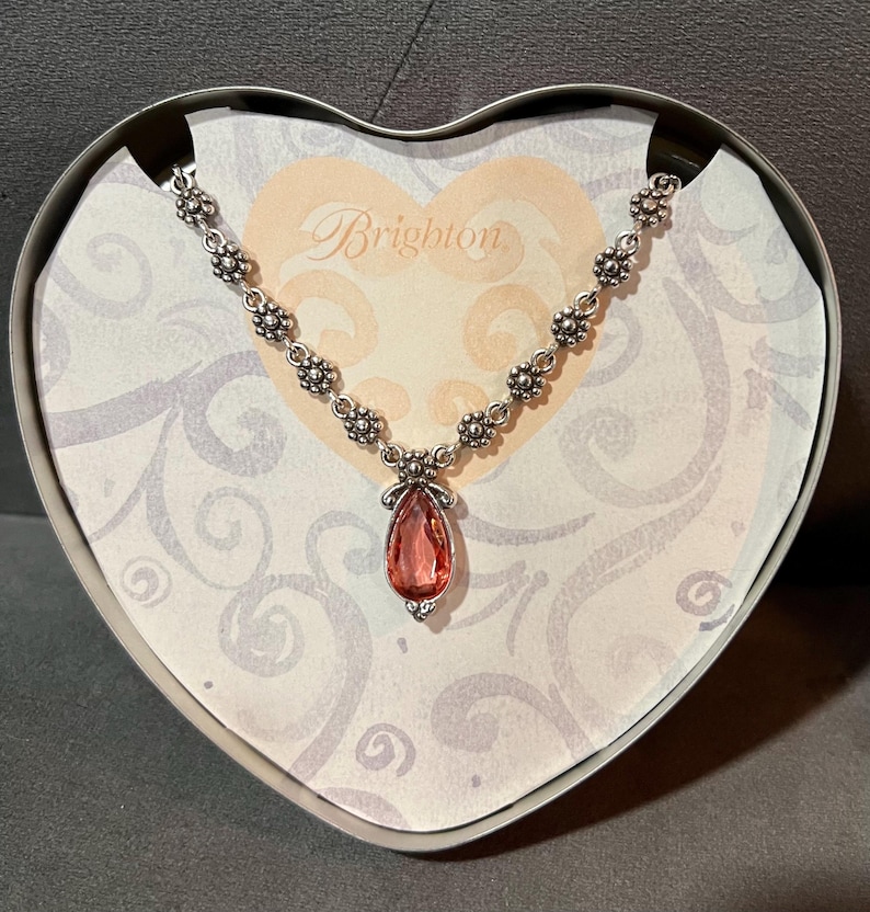May include: A silver-toned necklace with a teardrop-shaped, pink gemstone pendant. The necklace is displayed in a heart-shaped box with the brand name "Brighton" printed on the inside. The necklace has a floral design.
