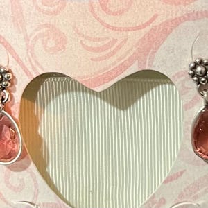 May include: A pair of silver-toned drop earrings with a floral design at the top and a teardrop-shaped, pink gemstone. The earrings are displayed in a heart-shaped box with a pink and white floral pattern.