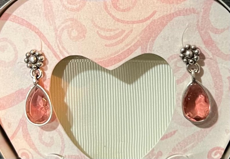 May include: A pair of silver-toned drop earrings with a floral design at the top and a teardrop-shaped, pink gemstone. The earrings are displayed in a heart-shaped box with a pink and white floral pattern.