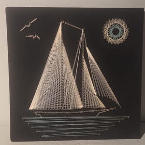 May include: A string art sailboat on a black background. The sailboat has white sails and is depicted with white string. Two birds and a sun are also made of string. The artwork is mounted on a square black canvas.