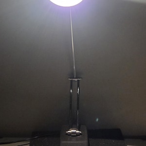 May include: A black desk lamp with a round, white light. The lamp has a silver, adjustable arm and a black base. The base has the text "TRANSFORMER POWERED" and the number "1". The lamp is on a black surface.