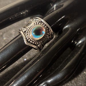 May include: A silver-toned ring with an iridescent, oval-shaped stone. The ring features ornate detailing around the stone and on the band. The ring is displayed on a black surface.
