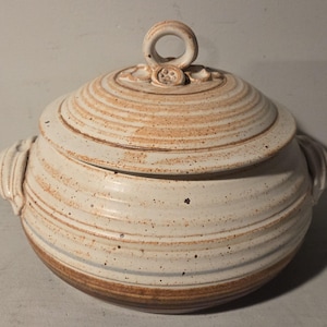 May include: A handcrafted ceramic lidded bowl with a cream and brown color scheme. The bowl features a round shape with horizontal ridges and decorative handles. The lid has a ring handle and floral accents. The pottery is speckled with dark spots.