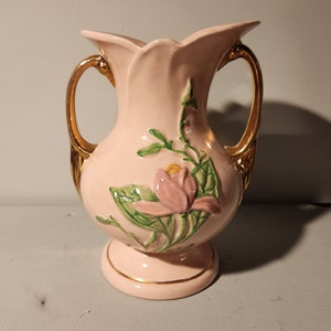 May include: A pink ceramic vase with gold handles and a floral design. The vase features a pink flower with green leaves.