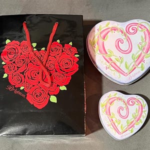 May include: A black gift bag with a red rose heart design and the word "Brighton". Two heart-shaped tins with pink heart designs and green vines. The tins have the word "Brighton" on them.