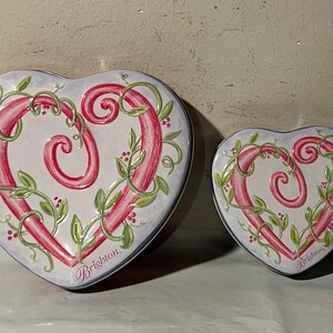 May include: Two heart-shaped decorative tins. The tins are white with a pink heart design, green vines, and red berries. The word "Brighton" is written on the bottom of each tin.