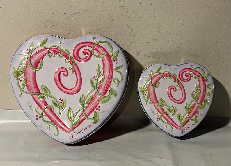 May include: Two heart-shaped decorative tins. The tins are white with a pink heart design, green vines, and red berries. The word "Brighton" is written on the bottom of each tin.
