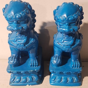 May include: A pair of blue ceramic foo dog statues. The statues are sitting on square bases and have intricate details on their bodies and faces.