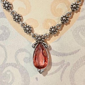 May include: Silver-toned necklace featuring a teardrop-shaped, pink gemstone pendant. The pendant is accented with decorative silver filigree. The necklace chain has a floral design. The jewelry is displayed on a patterned background.