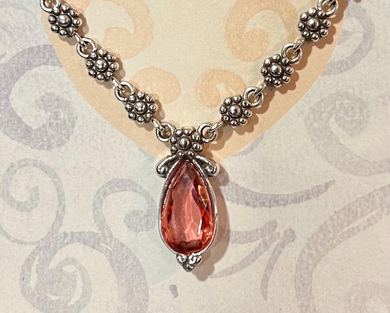 May include: Silver-toned necklace featuring a teardrop-shaped, pink gemstone pendant. The pendant is accented with decorative silver filigree. The necklace chain has a floral design. The jewelry is displayed on a patterned background.