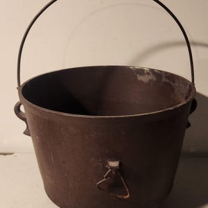 May include: A cast iron pot with a wire handle and a small metal loop on the side. The pot is dark brown and has a rustic appearance.