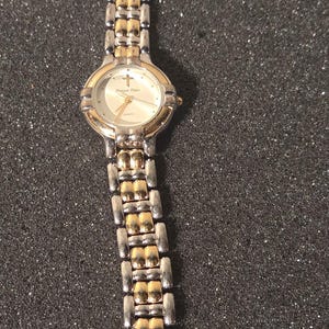 May include: A gold and silver-toned wristwatch with a rectangular clasp. The watch face is white with gold accents and a cross symbol. The watch band is a combination of gold and silver links. The watch reads "Prayer Time".