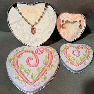 May include: A heart-shaped jewelry set featuring a necklace and earrings with pink gemstones, displayed in heart-shaped boxes. The boxes are decorated with floral designs and the brand name "Brighton".
