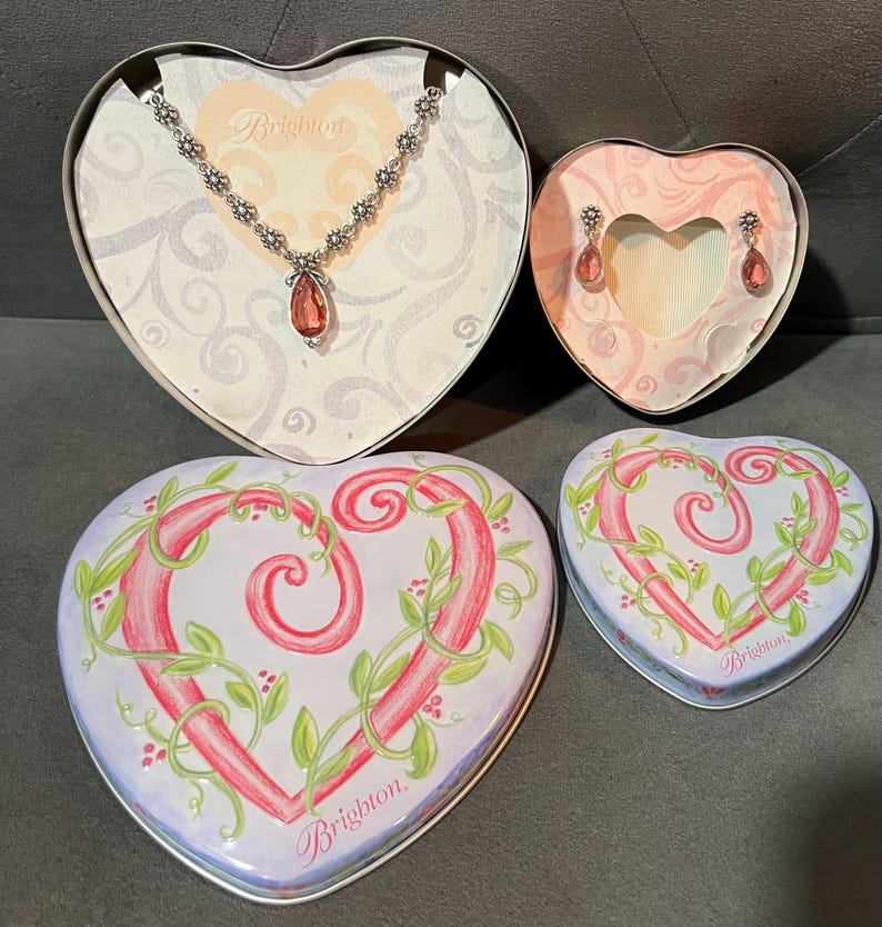 May include: A heart-shaped jewelry set featuring a necklace and earrings with pink gemstones, displayed in heart-shaped boxes. The boxes are decorated with floral designs and the brand name "Brighton".