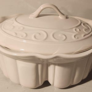 May include: White ceramic casserole dish with a lid. The dish has a scalloped edge and a decorative swirl design. The lid has a knob handle.