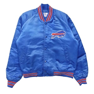 May include: A bright blue satin varsity jacket featuring a Buffalo Bills logo patch on the chest. The jacket has a snap-button front closure, two side pockets, and ribbed knit cuffs and hem with red and white stripes. The collar is also ribbed.