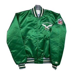 Puede incluir: Una chaqueta universitaria de satén verde con el logotipo de los Philadelphia Eagles bordado en el pecho, cuello y puños a rayas blancas y verdes, cierre de botones a presión, un parche de la NFL en la manga izquierda y logo de Starter.