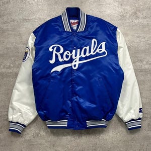 May include: A royal blue satin varsity jacket featuring white sleeves and the word Royals in white script across the chest. The jacket includes a striped collar, cuffs, and hem in blue, white, and grey, with a team logo patch on the right sleeve.
