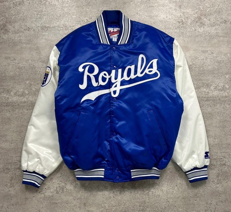 May include: A royal blue satin varsity jacket featuring white sleeves and the word Royals in white script across the chest. The jacket includes a striped collar, cuffs, and hem in blue, white, and grey, with a team logo patch on the right sleeve.