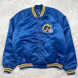 Puede incluir: Una chaqueta universitaria de satén azul real con cuello, puños y dobladillo acanalados a rayas amarillas y blancas. La parte delantera presenta un logotipo bordado de casco de fútbol americano Rams en blanco, amarillo y azul con botones.