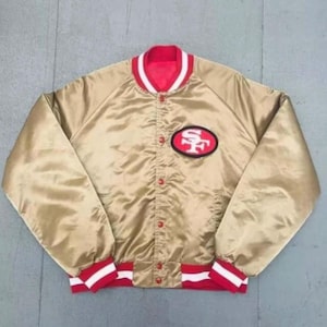 May include: A gold satin bomber jacket featuring a red and white striped collar and cuffs. The jacket has a front snap-button closure and a red and white oval patch on the left chest displaying the letters SF in a stylised font design.