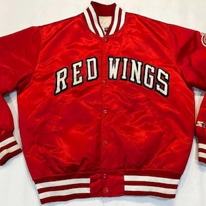 May include: A red satin jacket featuring the text "RED WINGS" in white block letters. The jacket has white and black striped trim on the collar, cuffs, and waist. A logo patch is visible on the upper left sleeve.