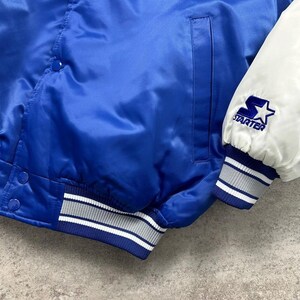 May include: A royal blue satin varsity jacket featuring white sleeves and a white Starter logo embroidered on the left cuff. The jacket includes a front snap button closure, a vertical side pocket, and ribbed knit cuffs and hem with white stripes.