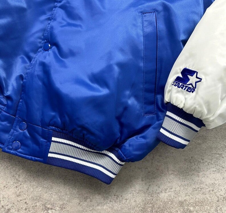 May include: A royal blue satin varsity jacket featuring white sleeves and a white Starter logo embroidered on the left cuff. The jacket includes a front snap button closure, a vertical side pocket, and ribbed knit cuffs and hem with white stripes.