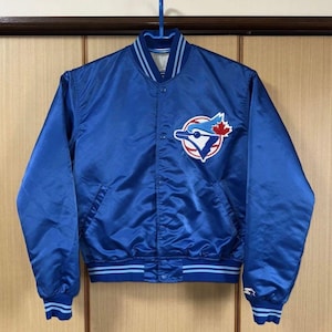 May include: A royal blue satin baseball jacket with a button-up front, featuring a team logo patch on the chest. The jacket has light blue and white striped accents on the collar, cuffs, and hem. The jacket is hanging on a blue hanger.